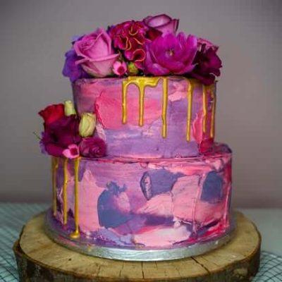 Tweelaagse stapeltaart met roze en paarse decoratie en bloemen, ontworpen door Bake My Day Groningen.